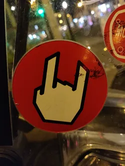 Street sticker A red circular sticker with a simplified, almost pixelated, white hand gesture signifying 'rock on'. The sticker shows some minor wear and tear, adding to its street aesthetic.