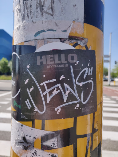 A black and white sticker with graffiti-style lettering. The sticker is affixed to a yellow pole, and other stickers are visible around it.