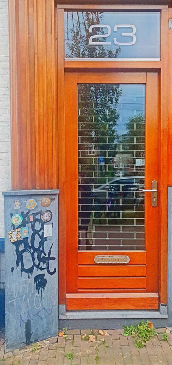 The image shows a corner pillar next to a wooden door. The pillar is covered in a variety of stickers and black graffiti. The wooden door has a metal grating over the glass and the number 23 above the door.