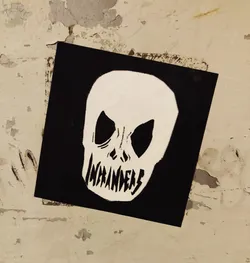 Street sticker Black and white sticker featuring a skull graphic with the word "Invaders" written across the lower jaw. The sticker has a dark background and is affixed to a textured wall.