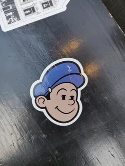 A vinyl sticker featuring a smiling cartoon sailor boy with a blue cap.  The style is reminiscent of classic cartoon characters. The sticker is affixed to a dark surface.
