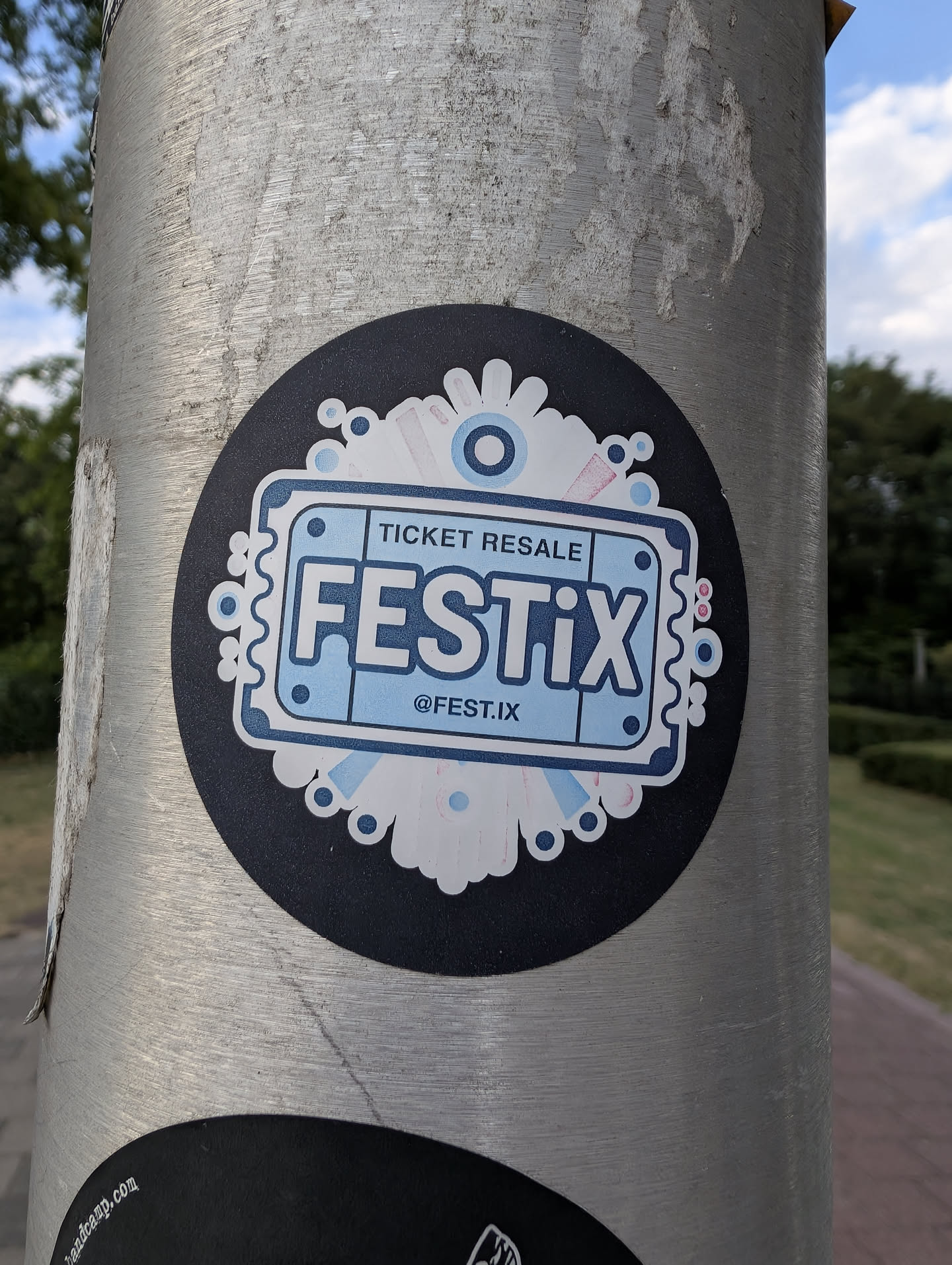 A round black sticker featuring a stylized light blue ticket with the text 'FESTIX' and 'TICKET RESALE'. The ticket is framed by a white, decorative floral pattern with blue and pink details.