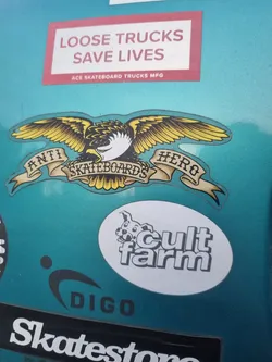 Street sticker A teal surface with various skateboarding brand stickers. The main focus is an Anti Hero Skateboards sticker featuring their iconic eagle logo with the words "Anti Hero Skateboards" written on a banner underneath the eagle. Other stickers include "Loose Trucks Save Lives", "Cult Farm", and "Digo".