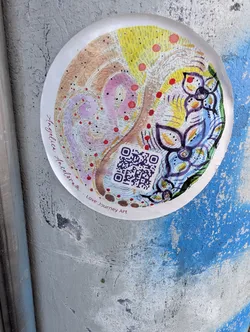 A round sticker featuring a colorful, hand-drawn abstract illustration with pastel swirls, yellow textures, purple flowers, and red dots. A QR code sits near the bottom, flanked by the text 'Angelica Angelorum - Love Journey Art'.