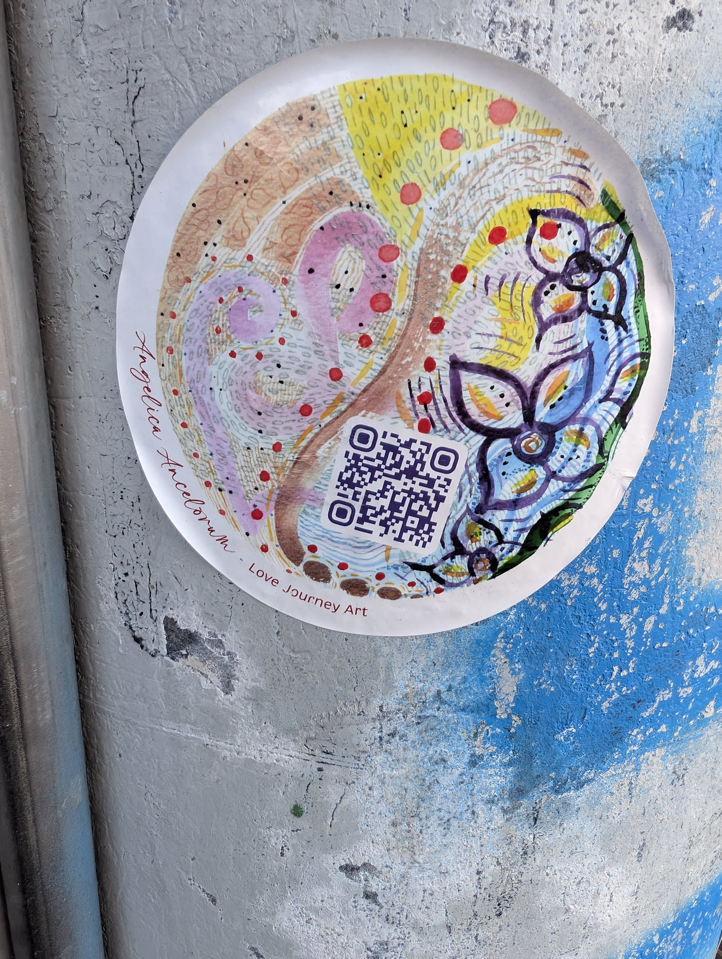 A round sticker featuring a colorful, hand-drawn abstract illustration with pastel swirls, yellow textures, purple flowers, and red dots. A QR code sits near the bottom, flanked by the text 'Angelica Angelorum - Love Journey Art'.