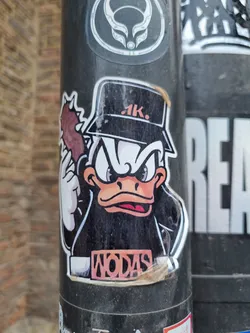 Street sticker A sticker featuring a cartoon duck wearing a cap and a leather jacket, holding what looks like a club. The duck has an angry expression. The text \"WODAS\" is visible on its jacket. The sticker is worn and slightly damaged around the edges. 