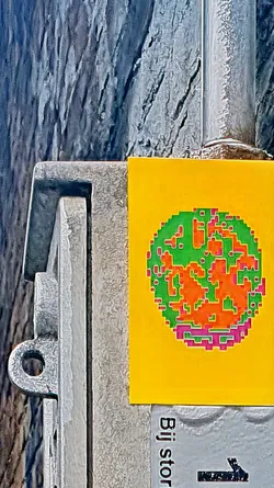 A sticker featuring a pixelated world map in green, orange, and pink against a vibrant yellow background is affixed to a metal pole. The map design is reminiscent of 8-bit graphics. The pole is textured, suggesting it's exposed to the elements. Part of another sign with the text "Bij stor" is visible below the sticker.