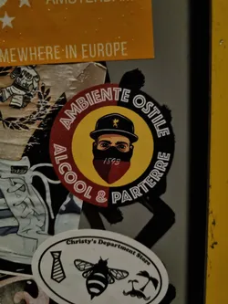 A round sticker with a yellow and red background. It features a black and white illustration of a person wearing a hat and a mask, with text surrounding it. The text says "AMBIENTE OSTILE" on the top and "ALCOOL & PARTERRE" on the bottom. The number 1893 is visible below the illustration.