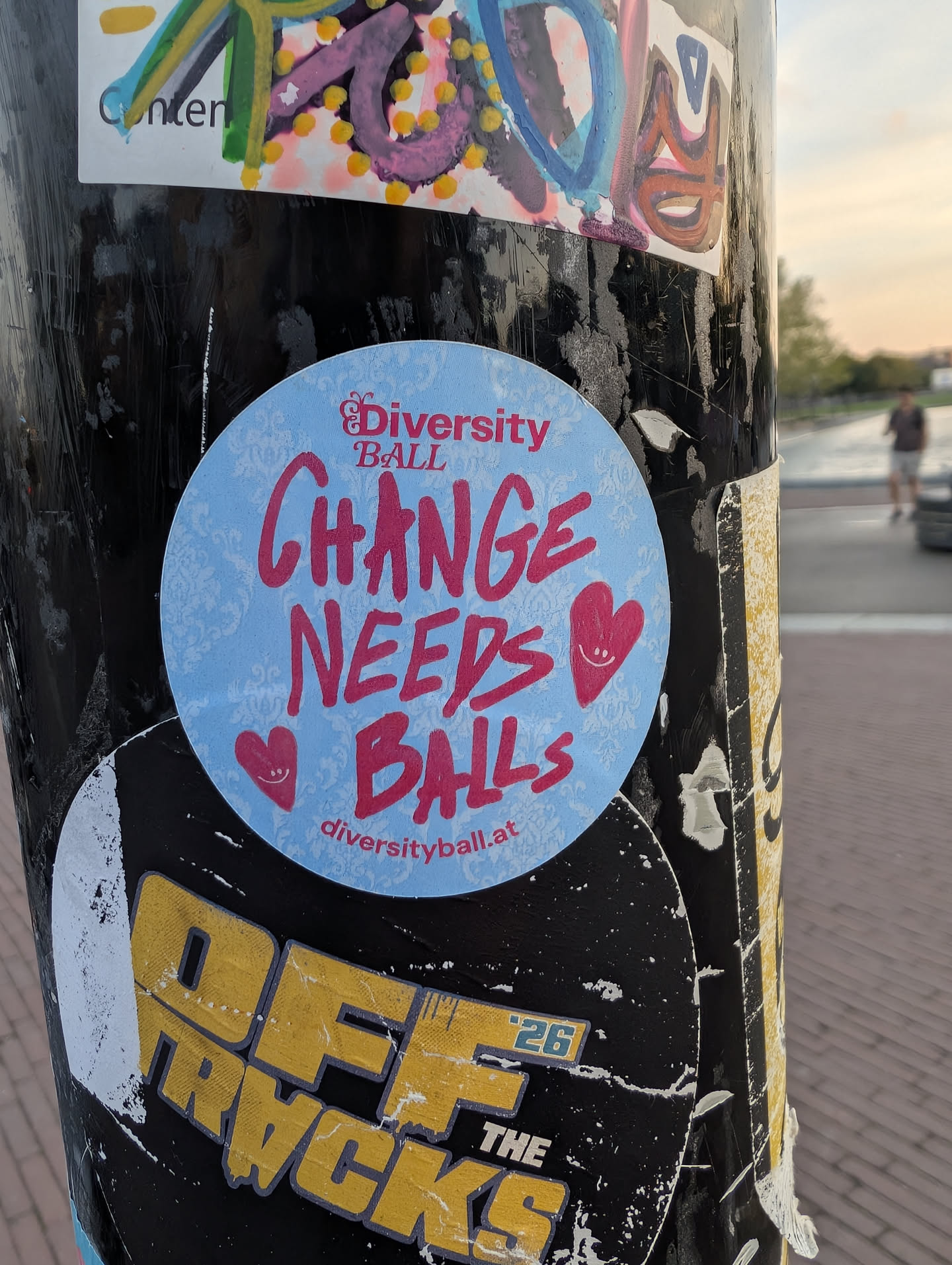 A round sticker with a light blue damask-patterned background. It features the pink text 'Diversity BALL' and 'CHANGE NEEDS BALLS' in a hand-drawn font, flanked by two smiling pink hearts. The URL 'diversityball.at' is printed at the bottom.