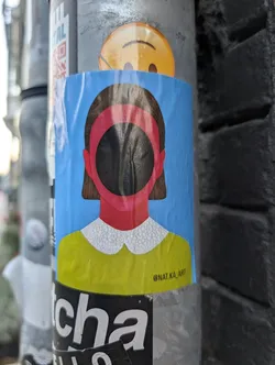 Street sticker A rectangular sticker depicting a stylized, faceless figure with brown hair against a blue background. The face is a black circular void ringed in pink. The figure wears a yellow top with a white patterned collar. The text '@NAT.KA_ART' is in the bottom right corner.