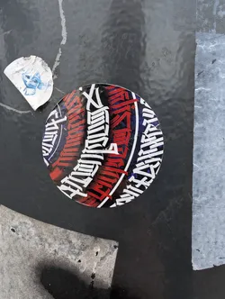 Street sticker A round sticker with illegible text in a stylized font. The text is primarily red and white, set against a black circular background. The sticker is affixed to a dark gray surface.