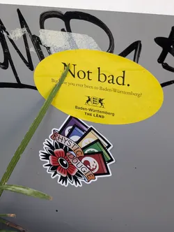 Street sticker An oval yellow sticker on a grey surface. It features the black text 'Not bad. But have you ever been to Baden-Württemberg?' alongside the state's coat of arms and 'THE LÄND' branding. A blade of grass crosses the left side.