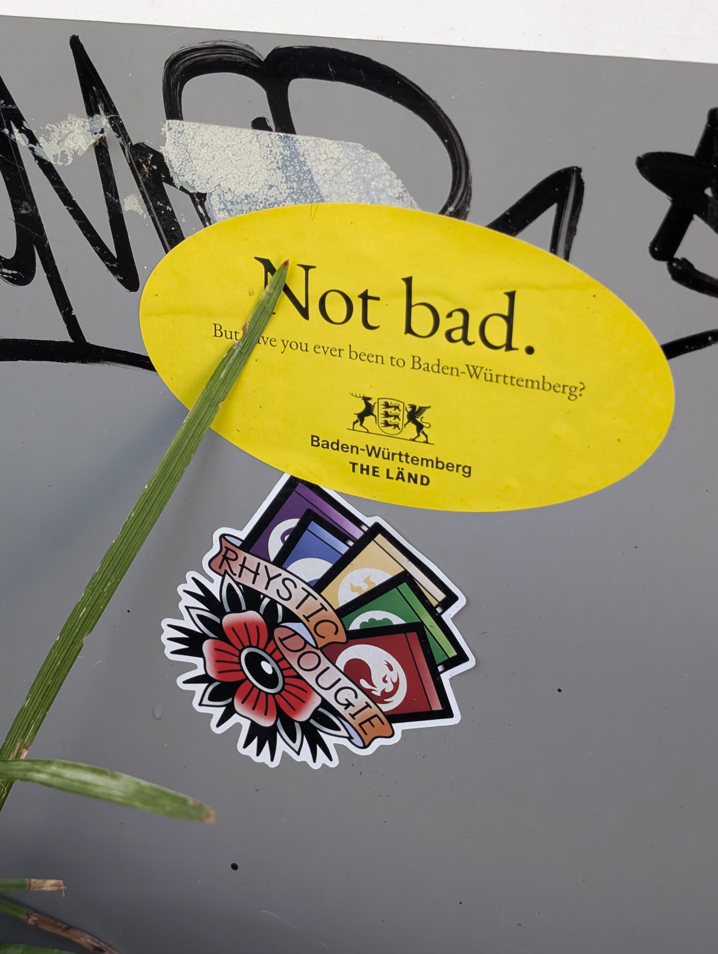 An oval yellow sticker on a grey surface. It features the black text 'Not bad. But have you ever been to Baden-Württemberg?' alongside the state's coat of arms and 'THE LÄND' branding. A blade of grass crosses the left side.