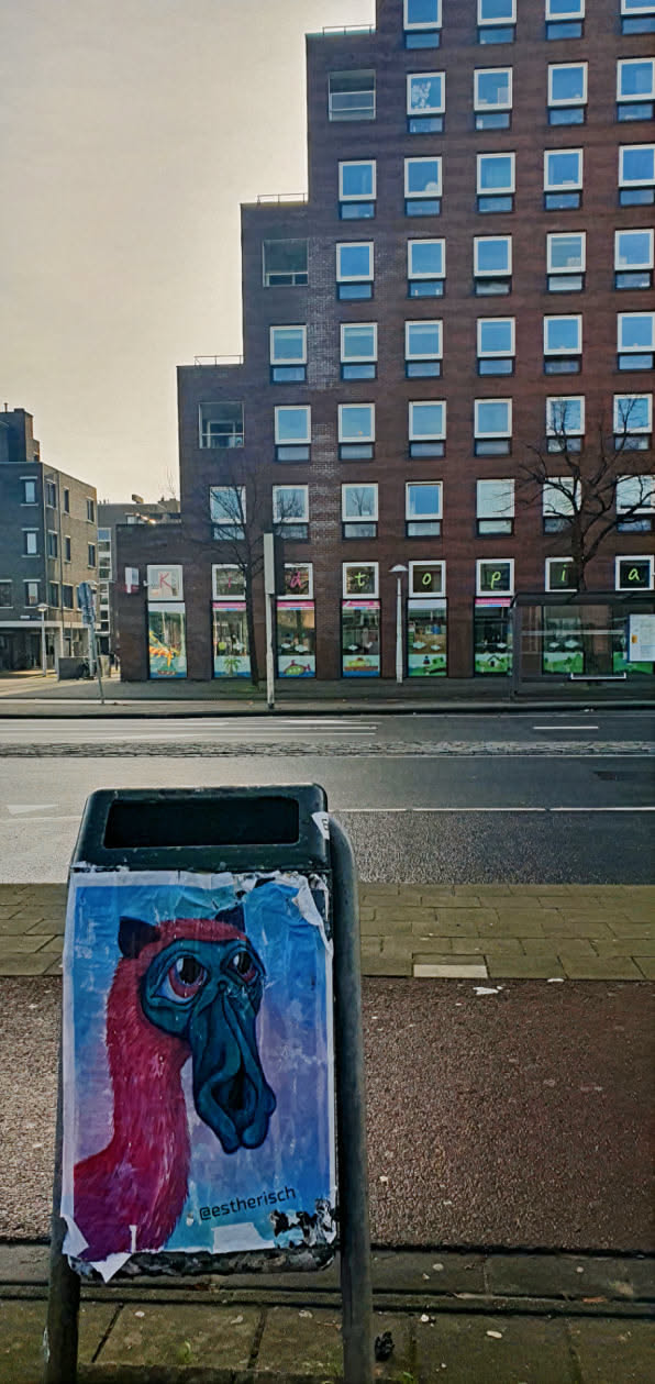 A colorful sticker featuring an illustration of a creature with a long snout and large, expressive eyes. The artwork is pasted on a trash bin, adding a touch of whimsy to the urban landscape. The sticker is torn at the edges.