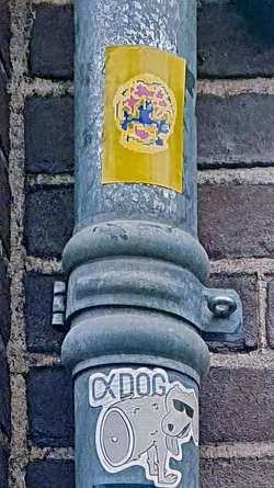 The image features a sticker adhered to a gray metal pole, which is positioned against a brick wall. The sticker has a yellow background with an abstract design in the center, featuring various geometric shapes and colors, including pink, blue, and yellow. Below the sticker is another sticker with a cartoonish dog character and text that reads "CxDog".