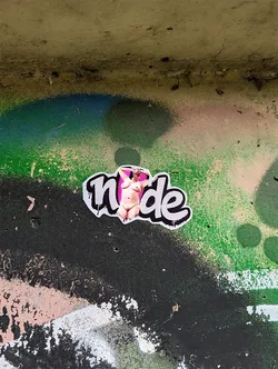 Street sticker Die-cut sticker spelling 'nude' in bubble font. The letters 'n', 'd', and 'e' are white with black outlines. The 'u' is replaced by a photographic cutout of a nude woman standing against a pink backdrop.