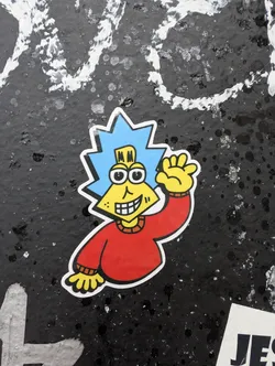 Street sticker The sticker features a cartoon character with a bright yellow, triangular face, large round eyes with black pupils, and a wide grin showing white teeth. The character has short, spiky blue hair and is wearing a red long-sleeved shirt with a yellow collar. The left hand is raised in a waving gesture. The sticker has a white border with a thin black outline.
