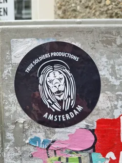 Street sticker A black and white round sticker featuring a lion with dreadlocks. The text 'TRUE SOLDIERS PRODUCTIONS' is written above the lion, and 'AMSTERDAM' is written below. The sticker is placed on a grey concrete surface.