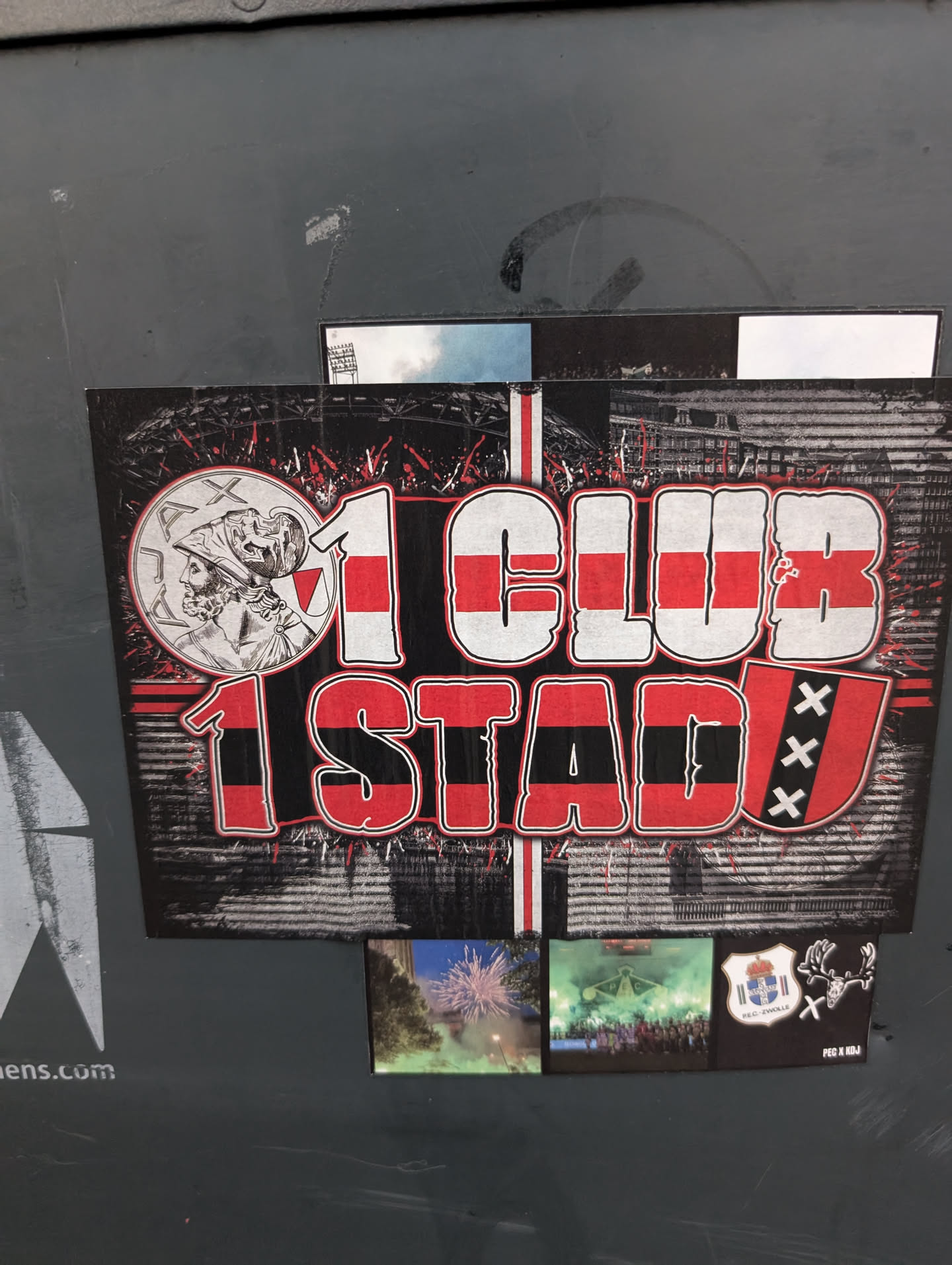 The central sticker is a large, rectangular graphic with a black and white distressed background depicting a stadium and crowd. Dominating the foreground are bold, stylized letters in red and white, spelling out '1 CLUB' above '1 STAD'. To the left of '1 CLUB' is a circular emblem featuring a profile of a classical figure wearing a helmet, possibly a Roman or Greek deity. To the right of '1 STAD' is a shield with three 'X' marks, representing the flag of Amsterdam. The overall design is energetic, with red paint splatters and a sense of movement. Below this main graphic are three smaller square images: one showing fireworks, another showing a crowd with green flares in a stadium, and a third with a coat of arms and antlers.
