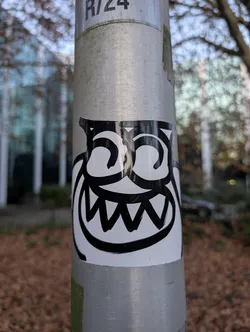 Street sticker The sticker features a stylized, black and white cartoon monster face. It has large, circular eyes with swirling pupils and a wide, jagged grin that resembles sharp teeth. The overall design is bold and graphic, with thick black outlines against a white background. The sticker appears to be adhered to a metal pole.
