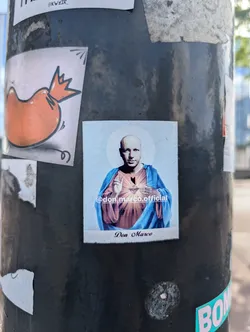 The sticker depicts a bald man dressed in robes, reminiscent of religious iconography. He has a halo around his head and is posed in a manner similar to depictions of Jesus or saints. The sticker appears to be an artistic or humorous take on religious imagery.