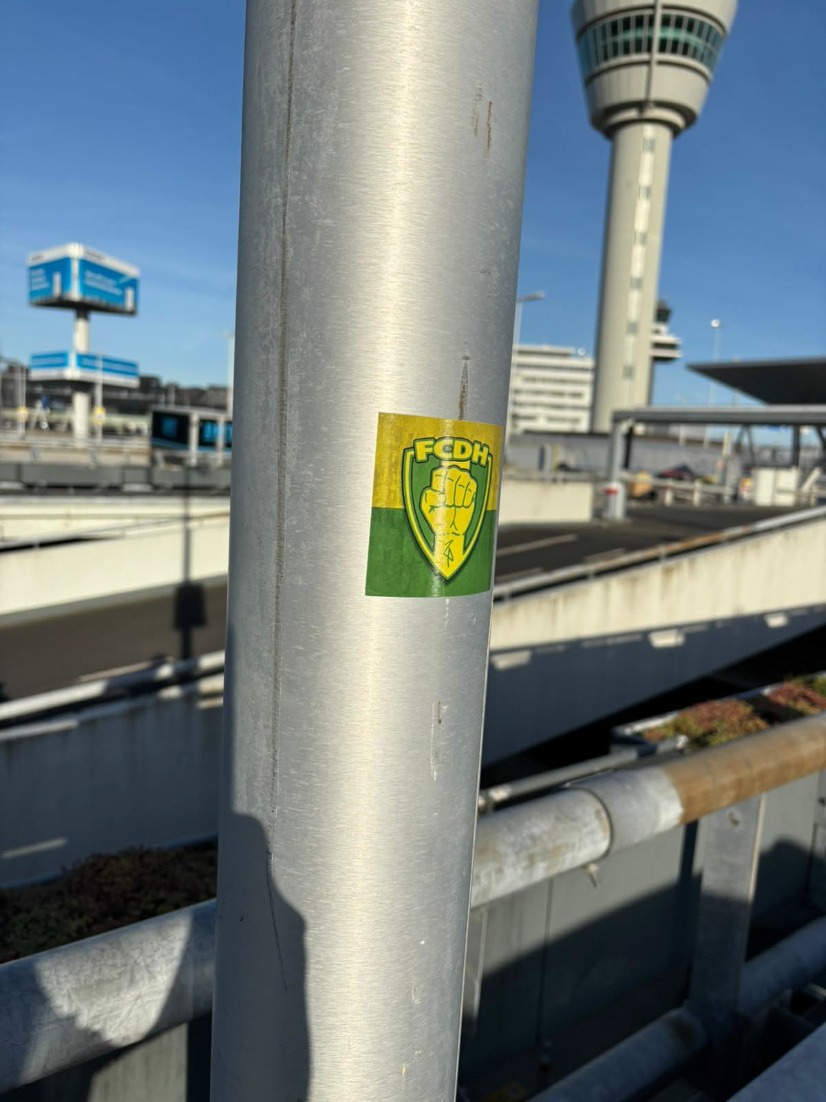 The sticker is rectangular with slightly rounded corners, featuring a split background of yellow on the top half and green on the bottom half. Centered on this background is a green shield outline, inside which a stylized yellow clenched fist is depicted, pointing upwards. Above the fist, on the yellow background, the letters "FCDH" are written in yellow. The sticker appears slightly worn and adhered to a metallic pole.