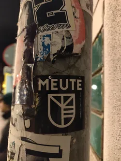 Black and white sticker with the word MEUTE and a logo that looks like a crest. The sticker is on a pole covered in other stickers.