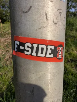 A rectangular sticker with a red background and white text that reads 'F-SIDE 76' is affixed to a metal pole. The number '76' is stylized within a circle with a checkered pattern, resembling a target.