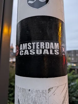 Street sticker A black and white sticker with the text "AMSTERDAM CASUALS" in a bold, stylized font.  There are small circular symbols on either side of the text.