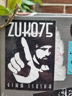 Black and white sticker featuring a stylized portrait of a man with a beard, making a number one hand gesture. The text "ZUKO75" is prominently displayed at the top, with smaller, less legible text at the bottom.