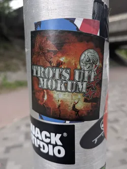 This sticker features the text "TROTS UIT MOKUM" in a stylized font, with a background evoking fire or explosions. Above the text is a helmeted figure, and below are silhouetted figures with arms raised. To the right is a coin-like image with a profile and red crosses. The overall design is somewhat edgy.