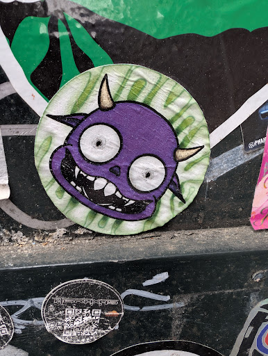 The sticker features a purple cartoon demon with small horns, wide eyes, and a toothy grin. It is circular with a green striped background.
