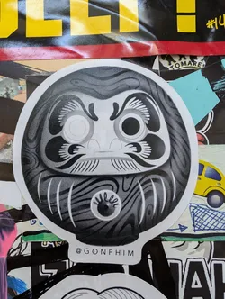 Street sticker Black and white illustration of a Daruma doll, a traditional Japanese good luck charm. The sticker has a stylized design with wood grain texture and a single eye. The artist's Instagram handle is printed at the bottom.