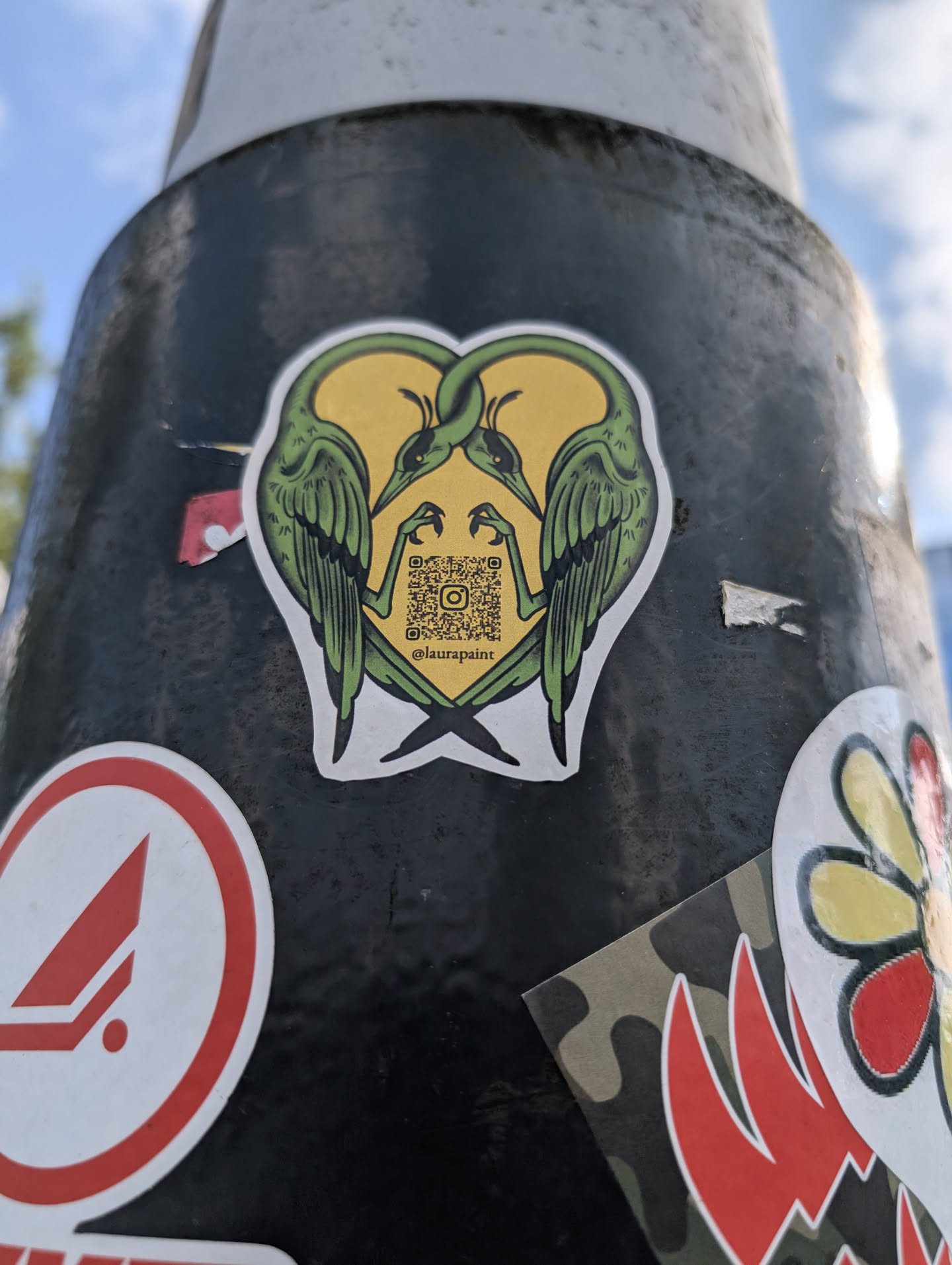 Street sticker This is a sticker of two green heron birds facing each other, forming a heart shape. The background is yellow, and there is a QR code with the Instagram username @laurapaint. The birds have a stylized design with detailed feathers and a striking pose.