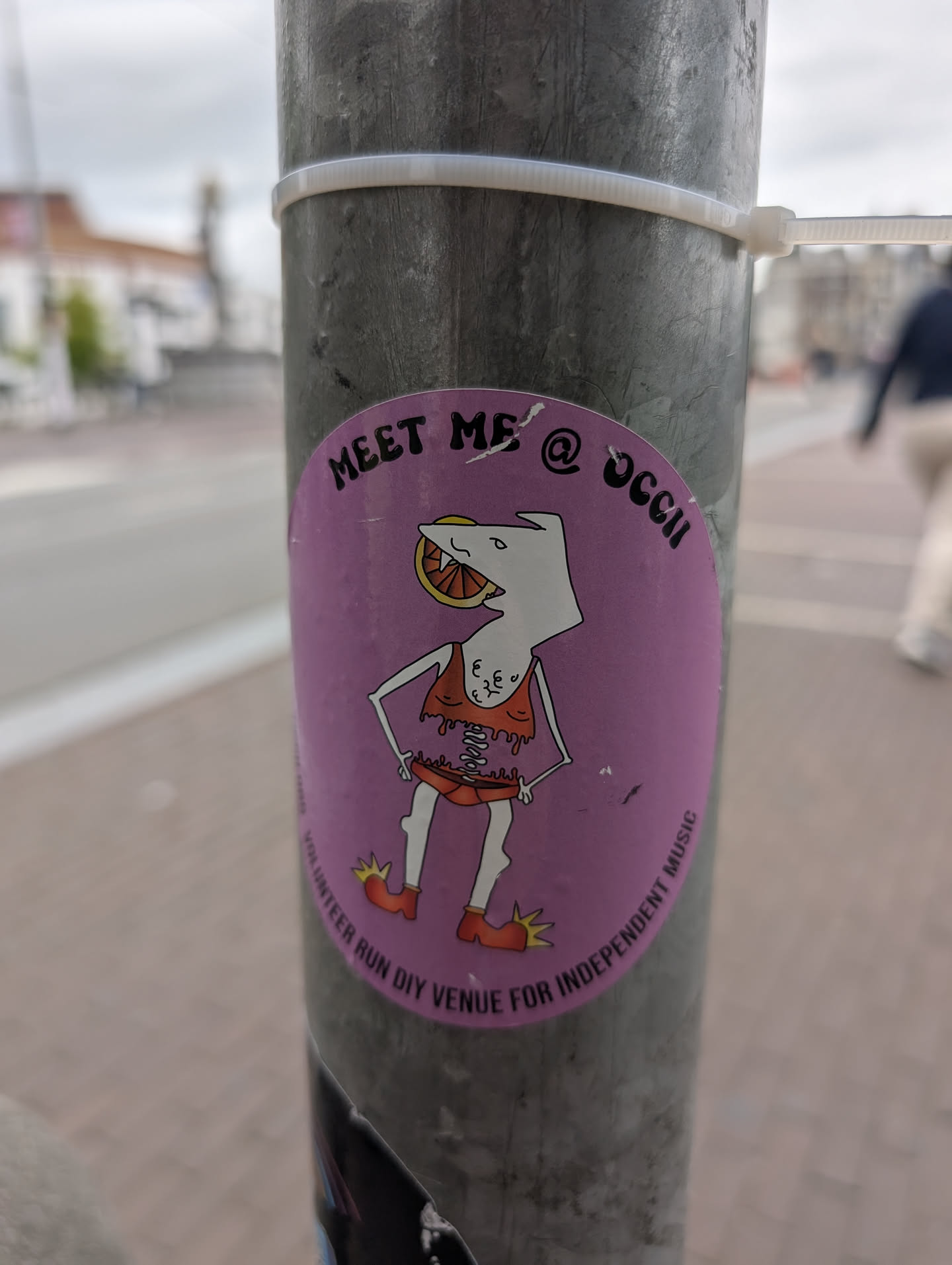 The sticker is oval-shaped with a vibrant pink background. In the center, there's a cartoonish character with a white head resembling a bird or a shark, with a slice of orange in its mouth. The character has a lanky, humanoid body with a bare, reddish-orange top with black lacing and ruffled edges, and short, orange shorts. It has thin, white legs with red, pointed shoes. The character's hands are on its hips in a confident pose. Below the character, curved text reads 'VOLUNTEER RUN DIY VENUE FOR INDEPENDENT MUSIC'. The top of the sticker features text in a stylized font: 'MEET ME @ OCCII'.