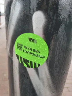Street sticker A round green sticker with a QR code and the text 'EGOless Expression'. The sticker has water droplets on it and is attached to a dark pole with white graffiti.  The logo 'KSII RECORDINGS' is also visible on the sticker.