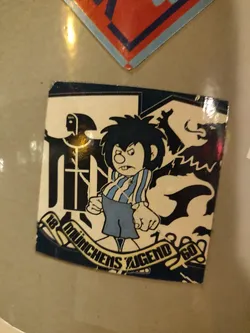 Street sticker A sticker featuring a cartoon character with spiky hair, wearing a striped shirt and shorts. The character has an angry expression. The sticker also features a banner with the text "18 Münchens Jugend 60" and some other graphical elements.