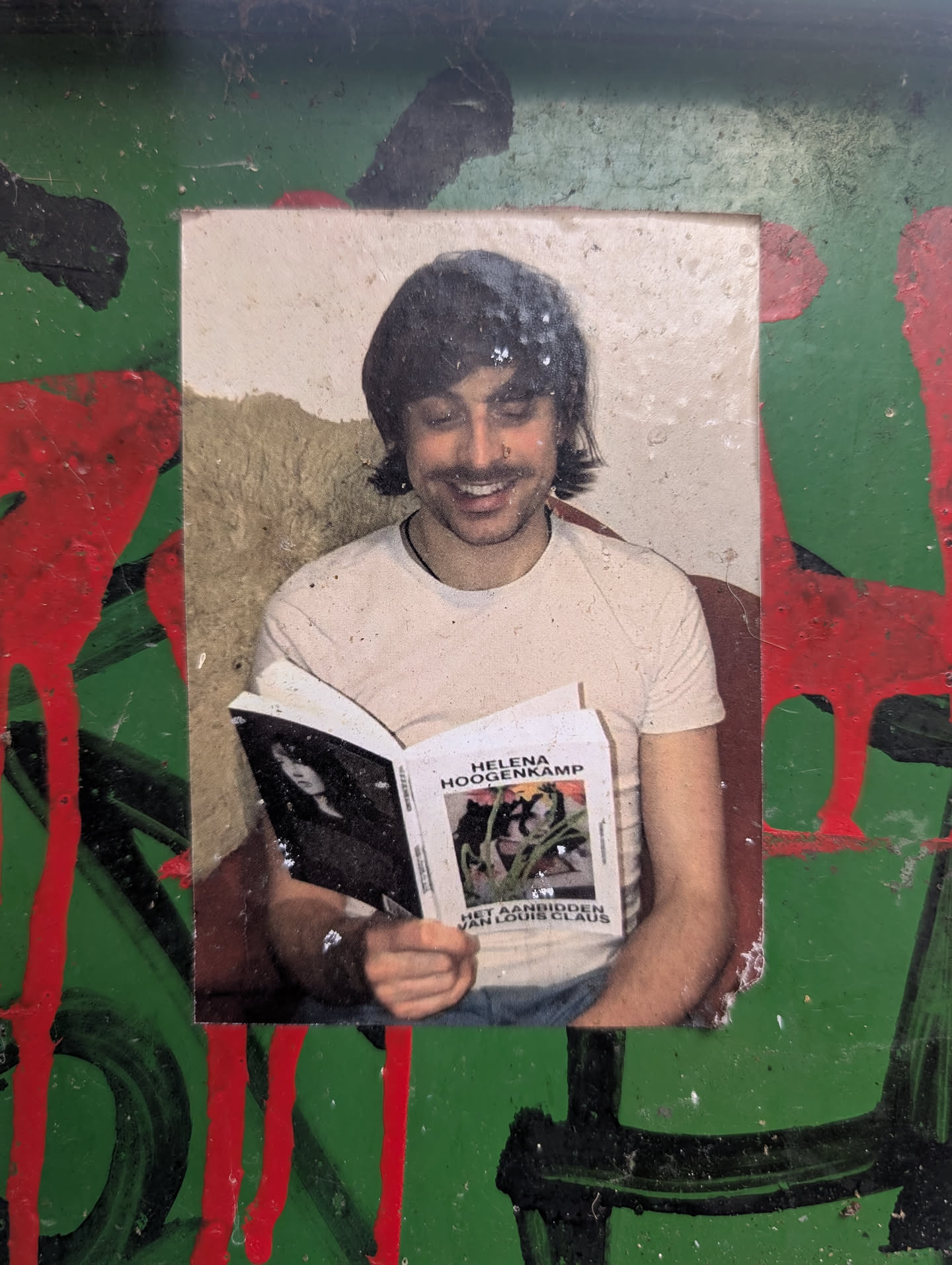 The sticker features a photograph of a smiling person with dark, wavy hair, wearing a light-colored t-shirt and blue jeans. The person is holding open a book. The book's cover shows a black and white image of a woman and text that reads 'HELENA HOOGENKAMP HET AANBIDDEN VAN LOUIS CLAUS'. The sticker is surrounded by a green background with abstract splatters and drips of red and black paint, giving it a street art or graffiti aesthetic.