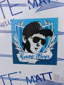 Street sticker A sticker featuring a stylized illustration of a young boy wearing a cap and sunglasses. The design is set against a blue background with a shield-like frame and laurel leaves. The text "Young Boyz" is prominently displayed in a stylized banner below the illustration.