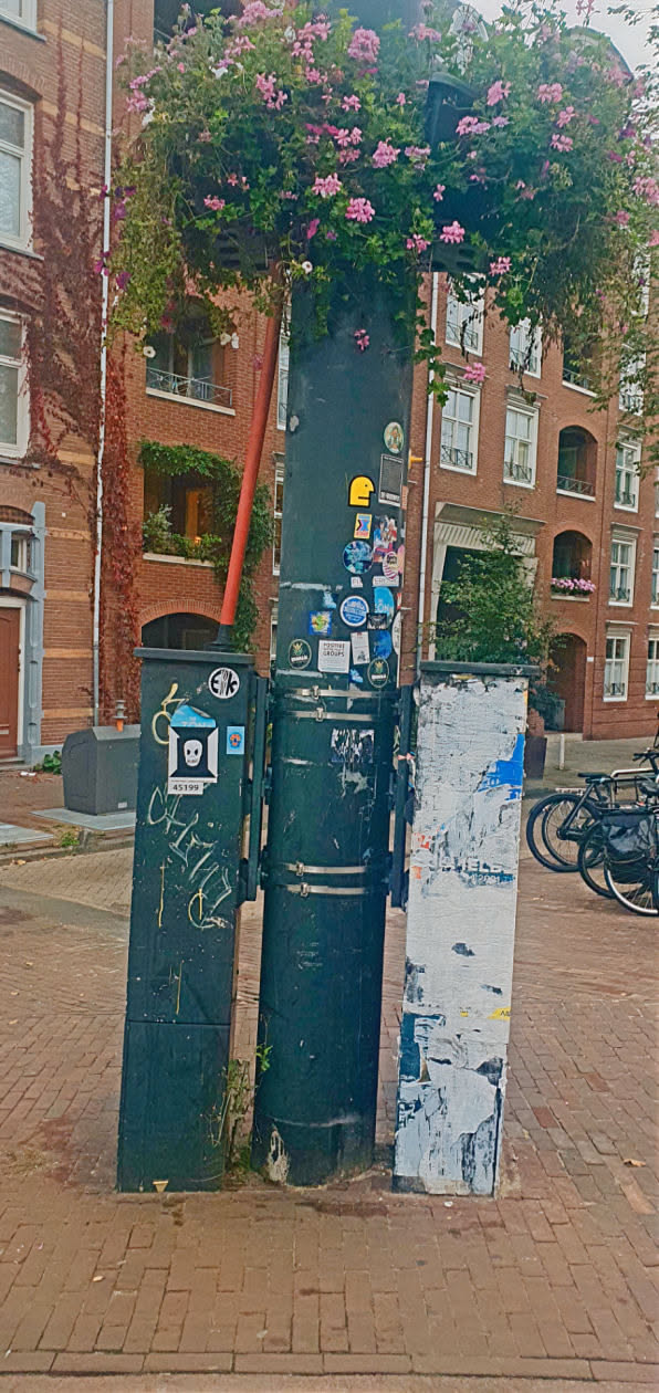 A street scene with stickers on a black pylon. Focus on the skull with a white frame sticker on the left pylon. There are other stickers and some graffiti near it.
