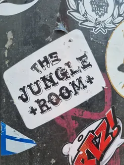 Close-up of a sticker featuring the text "THE JUNGLE ROOM +" in a stylized font. The sticker is white with black text and appears to be affixed to a surface with other stickers.
