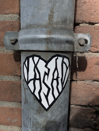 A white heart-shaped sticker with a black outline is placed on a gray metal pipe. Inside the heart, the text 'YASAY' is written in a stylized, fragmented font. The pipe is attached to a brick wall.