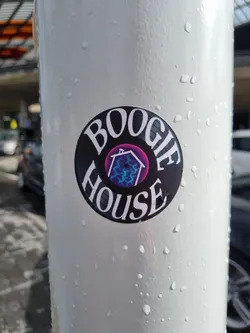 Street sticker A round sticker featuring the text 'BOOGIE HOUSE' in a stylized font reminiscent of vinyl records.  Inside the circle is a smaller image of a house with purple and blue lightning bolt designs. The sticker is affixed to a light gray pole outdoors, and there are water droplets visible on the pole's surface.