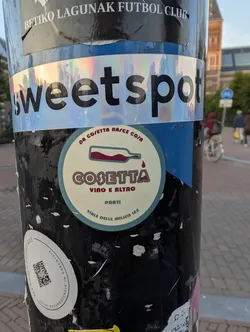 Street sticker A round, cream-colored sticker with a teal border. It features a minimalist illustration of a horizontal wine bottle pouring a single red drop. Below the bottle, the stylized word 'COSETTA' is printed in outlined letters, followed by 'VINO E ALTRO', 'PRATI', and the address 'VIALE DELLE MILIZIE 122'.