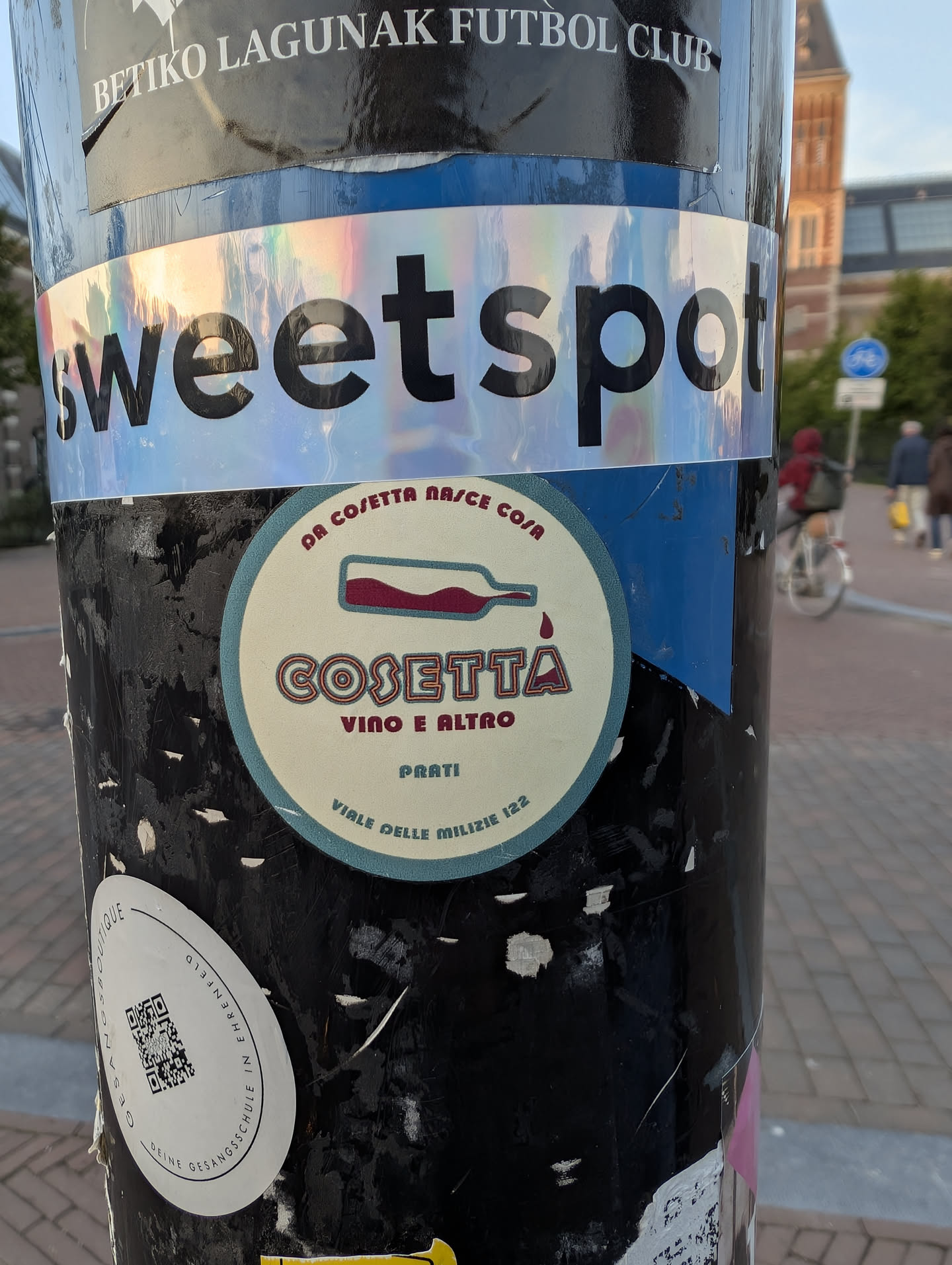 A round, cream-colored sticker with a teal border. It features a minimalist illustration of a horizontal wine bottle pouring a single red drop. Below the bottle, the stylized word 'COSETTA' is printed in outlined letters, followed by 'VINO E ALTRO', 'PRATI', and the address 'VIALE DELLE MILIZIE 122'.