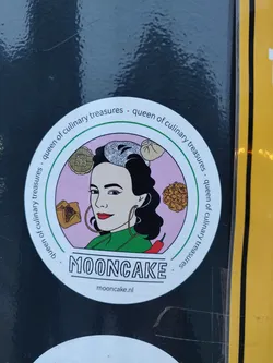 Street sticker A round sticker featuring a stylized illustration of a woman with dark hair, large hoop earrings, and a green collared shirt. She is surrounded by various Asian-inspired food items such as mooncakes, dumplings and what appears to be a chili pepper. The text 'MOONCAKE' is prominently displayed below the illustration with the website mooncake.nl underneath. The phrase 'Queen of Culinary Treasures' is written around the outer edge of the sticker.