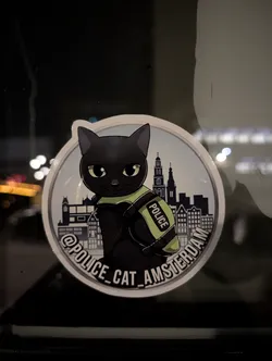 Street sticker A circular sticker featuring a stylized black cat with large, light green eyes, wearing a bright lime green police vest with 'POLICE' written vertically in white. Behind the cat is a silhouette of the Amsterdam skyline, including iconic canal houses and church spires, rendered in shades of grey and white against a light blue background. The sticker has a white outer border, and along the bottom edge, in a curved arc, is the text '@POLICE_CAT_AMSTERDAM' in white letters with a black outline. The overall aesthetic is cartoonish and charming.