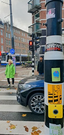 A black and white traffic pole in Amsterdam is adorned with several stickers. The pole is in front of a crosswalk where a woman in a green coat is crossing the street. The stickers include an orange one with a person pressing a button, a green one with a blue and red globe shape, and text at the bottom that says "Breathe In, Burn Out."
