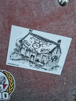 Black and white drawing of a house with a banner that says "Support your local squat".  There is an anarchy symbol on the house. The website zwartekat.org is printed at the bottom.