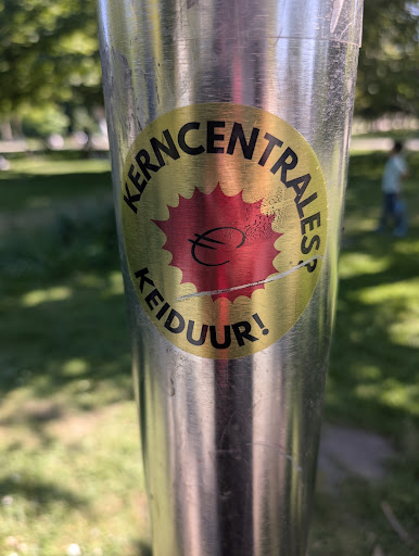 This sticker on a pole has a golden background with a red burst shape in the center, superimposed with a euro currency symbol. The text "KERNCENTRALES? KEIDUUR!" is printed around the edge.